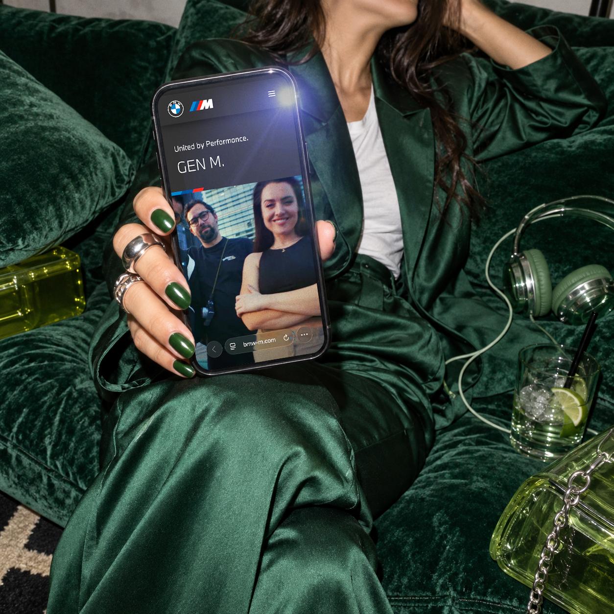 A person in a green satin outfit holds a smartphone featuring the BMW-M.com webpage.
